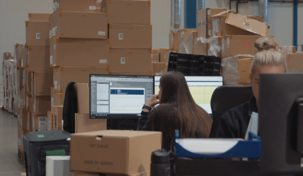 A picture of workers wokring at working stations with boxes in the background.