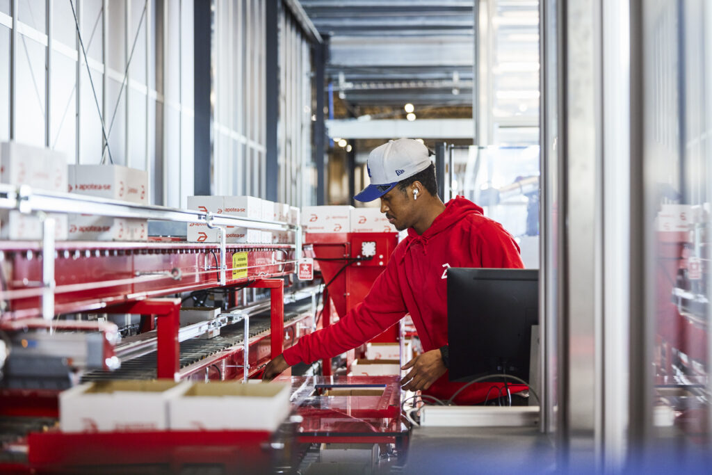 Operario trabaja en una estación automatizada de preparación de pedidos junto a un monitor y líneas de transporte de cajas.