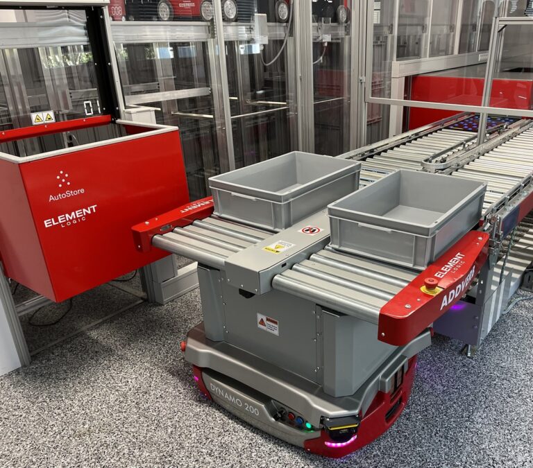 Robot móvil autónomo Dynamo 200 de Addverb transporta cubetas frente a una estación AutoStore y una línea de rodillos en una instalación de automatización intralogística de Element Logic.