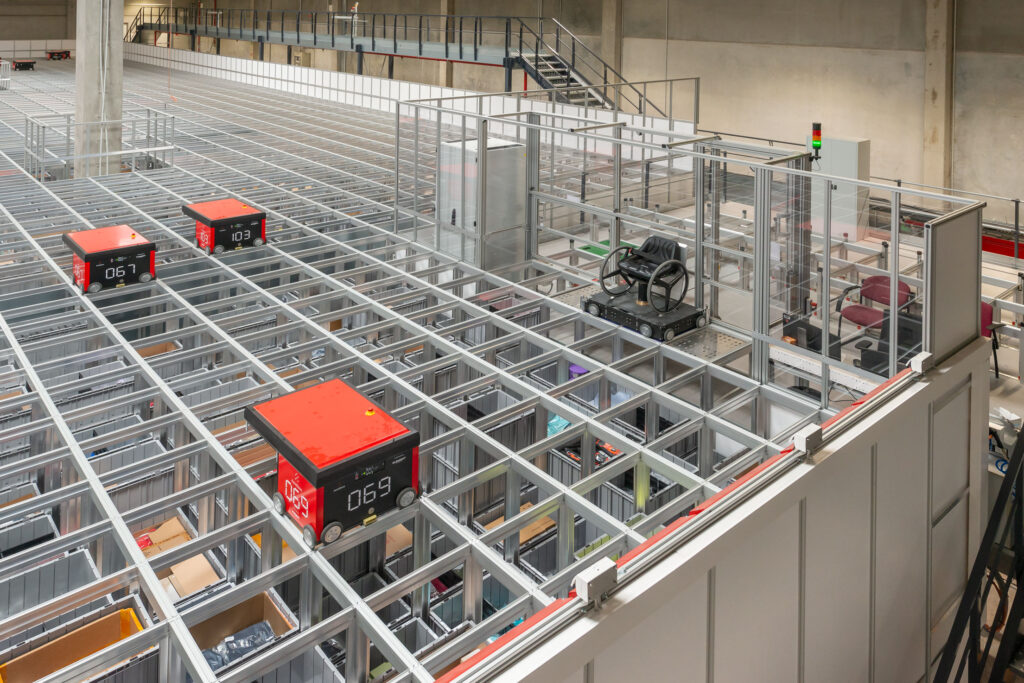 obots AutoStore se desplazan sobre una cuadrícula de almacenamiento junto a una estación adaptada para accesibilidad en un almacén automatizado.