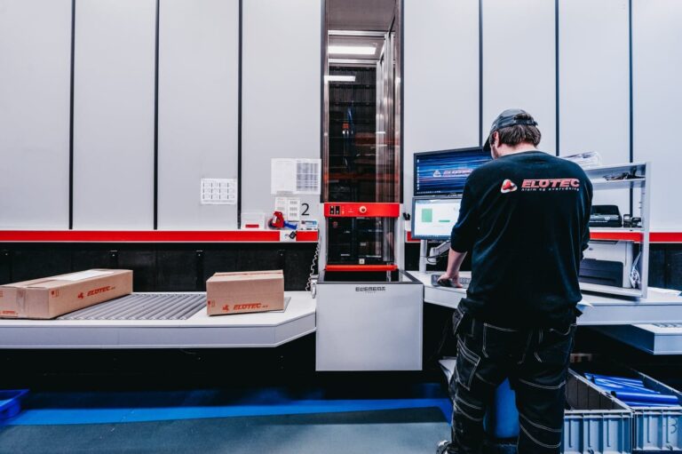 Operario trabaja en una estación AutoStore con cinta de rodillos, cajas y pantalla de control en un almacén automatizado.