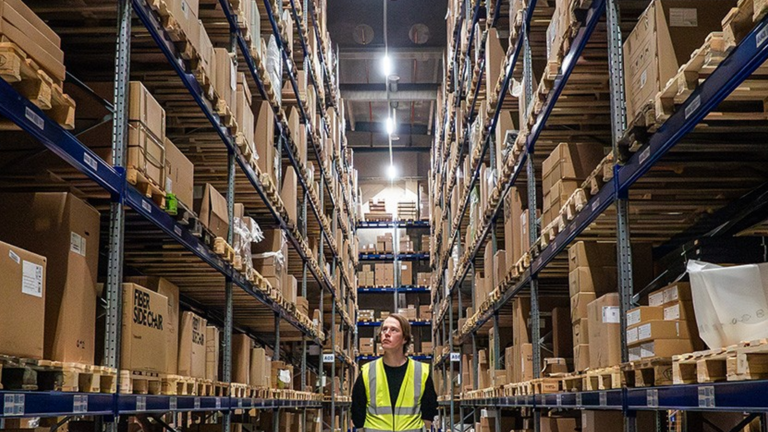 Operario con chaleco de alta visibilidad camina por un pasillo entre estanterías altas con cajas y palets en un almacén.