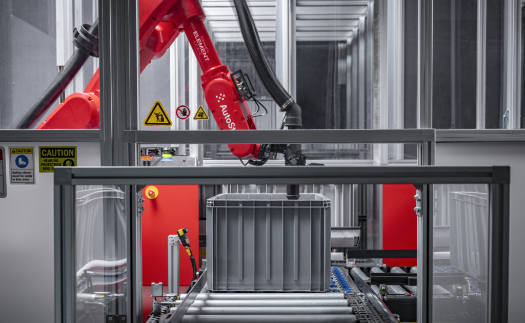 Brazo robótico de AutoStore con marca Element Logic manipulando una cubeta en una célula automatizada.