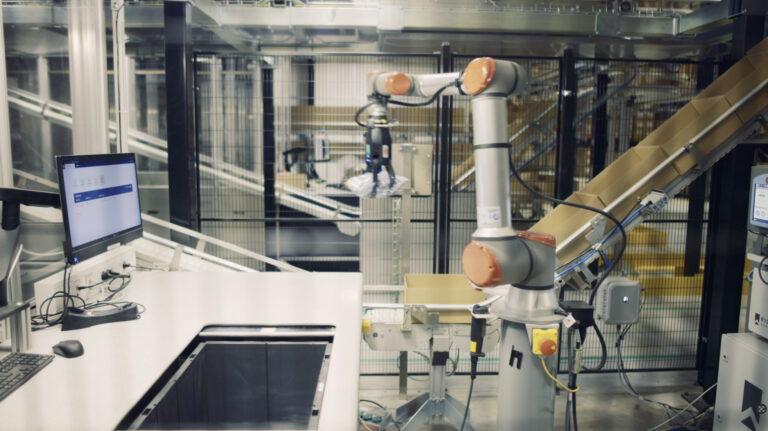 Estación de automatización de almacén con brazo robótico y puesto de trabajo con pantalla.