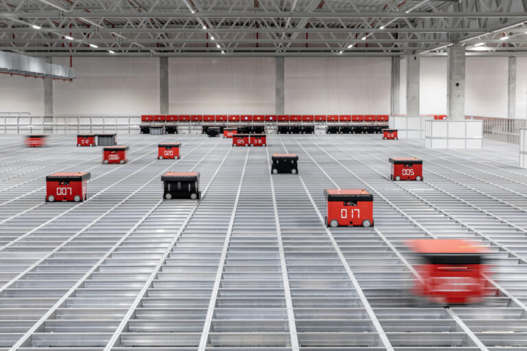 Robots móviles rojos y negros se desplazan sobre una gran estructura metálica en un almacén automatizado, con filas de estaciones al fondo y techo industrial iluminado.
