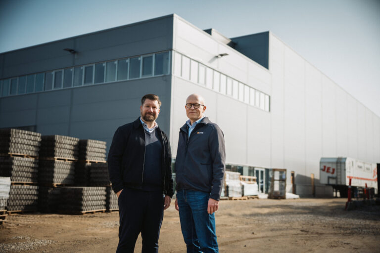 To mænd står foran et stort industrilager med paller og byggematerialer i baggrunden.